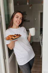 Pretty smiling woman in white t-shirt is holding plate with croissants and morning coffee while standing at kitchen with happy smile. Lovely woman is having breakfast in the morning at home