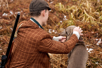 Naklejka premium rear view on caucasian young man with gray dog sitting in field waiting for target wild animal trophy, young guy in brown suit in nature, with shotgun. hunting, activ lifestyle, hobby concept