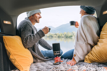 Car trunk view of chatting couple dressed warm knitted clothes enjoying gas stove prepared coffee and mountain lake view. Cozy early autumn couple auto traveling concept image..