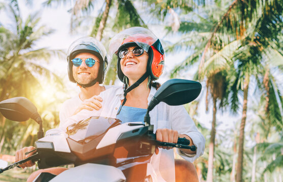 Happy smiling couple travelers riding motorbike scooter in safety helmets during tropical vacation under palm trees on Ko Samui island , Thailand