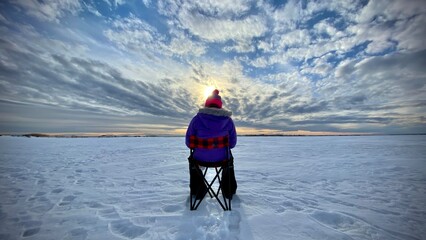 A person sitting in a chair ice fishing on a frozen lake in Newell County Alberta Canada under a dramatic sunset sky.
