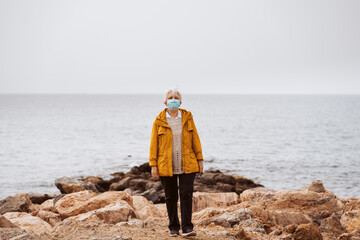 .Middle-aged woman enjoying the new normal in front of the sea, wearing a mask and using her smartphone.