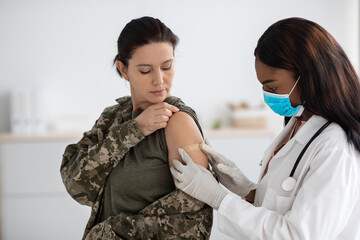 Black Female Doctor Applying Plaster On Soldier Woman's Shoulder After Injection Shot