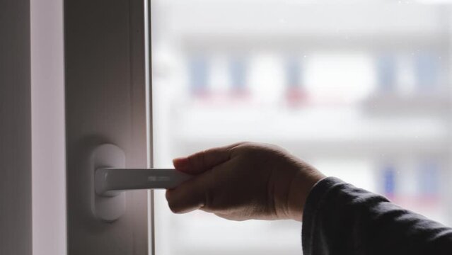 close-up of female hand behind plastic handle opens new modern window in an apartment of blurred multi-storey building, the concept of ventilating housing, fighting mold, fresh air, saving heat