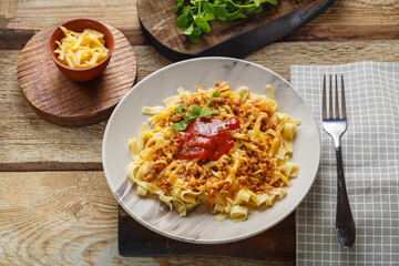 Pasta bolognese garnished with greens and cheese in a plate on a wooden table on a stand next to a fork and greens on carved boards.