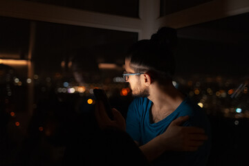 Night portrait of young man with smartphone in hand looking through window.