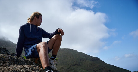 A bottom view of young man sitting on a rock in the mountains