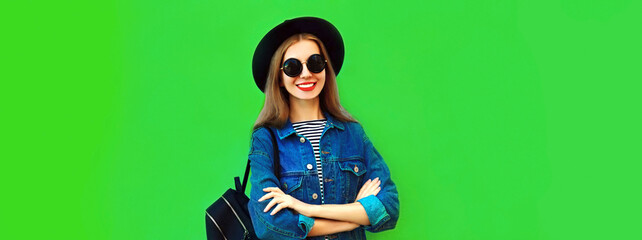 Portrait of smiling young woman model wearing black round hat, denim jacket and backpack on green...