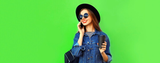 Portrait of happy smiling young woman calling on smartphone wearing black round hat, denim jacket on green background