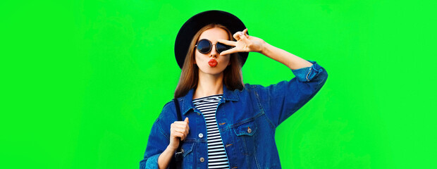 Portrait of stylish young woman blowing her red lips sending air kiss wearing black round hat, denim jacket on green background