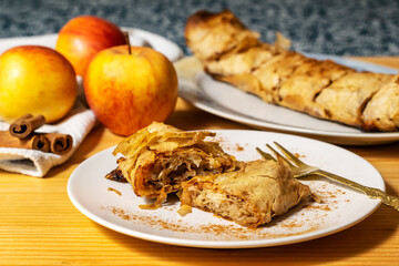 A roll of homemade baked fresh apple pie strudel with an appetizing slice of apple pie on the plate and fresh apples and cinnamon sticks on a wooden table. Rustic breakfast concept