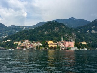 Varenna view, Italy. Colourful coastal houses view from Como lake. Hills and mountains. Forest, park, cloudy day, yellow buildings, bell tower.