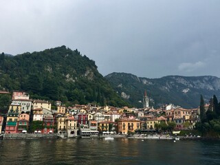Fototapeta na wymiar Varenna view, Italy. Colourful coastal houses view from Como lake. Hills and mountains. Forest, park, cloudy day, yellow buildings, bell tower.