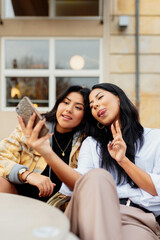 Two Latin female friends taking a selfie gesturing with their smartphone. Latin sisters having good times together in the street.