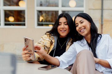 two hispanic sisters taking a picture with their smart phone in the street. Young latin girls using...