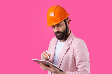 Portrait of young construction engineer wear orange hard hat, in a pink jacket standing on red studio background. A man with a tablet in his hands.