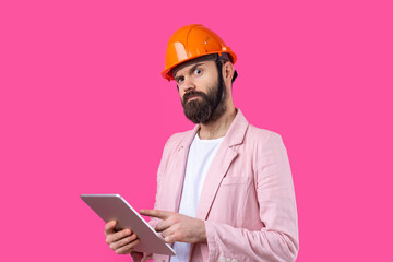 Portrait of young construction engineer wear orange hard hat, in a pink jacket standing on red studio background. A man with a tablet in his hands.