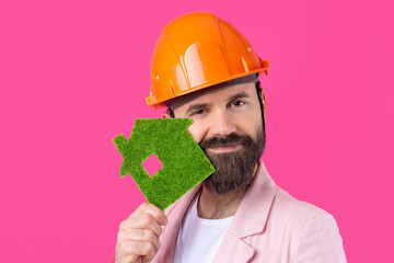 Portrait of young construction engineer wear orange hard hat, in a pink jacket standing on red studio background. A man holds a green eco house.