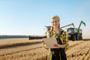 Farmer on grain field with tractor and combine harvester in background taking notes