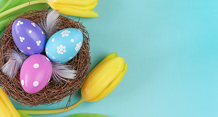 Easter eggs lie in a nest with yellow tulips arrangement on blue background. Flat lay top view with copy space. Happy Easter holiday.