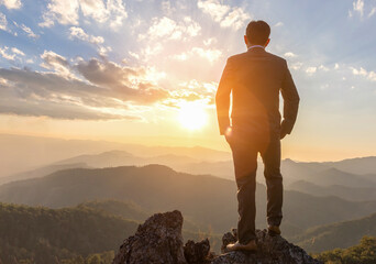 Successful businessman in business suit standing and looking sunset on top rock mountain background,  competition and leadership concept.