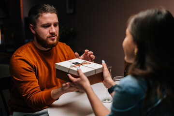 Happy young man receiving present from loving wife enjoying romantic dinner date sitting at table with candles on birthday or Valentines Day. Pretty young woman giving wrapped box with gift to husband