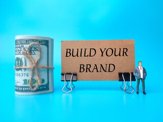 Banknotes and miniature people with text BUILD YOUR BRAND on blue background.