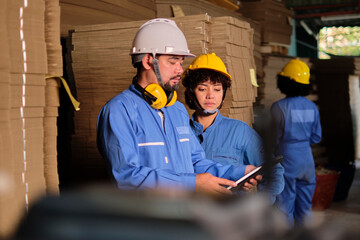 Asian male engineer in safety uniform and hard hat and female worker colleague inspect storage, stock order at factory warehouse, piles of stacking cardboard manufacture, industry product management.