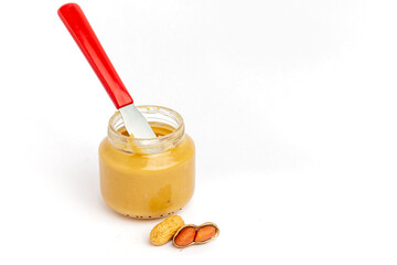 Glass jar with peanut butter, butter knife and peanuts in front on white background.