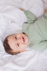Fototapeta na wymiar portrait of a cute smiling baby in a green cotton bodysuit lying on a white bed on his back