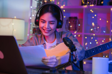 Cheerful beautiful Asian woman wearing headphones and playing acoustic guitar with using laptop...