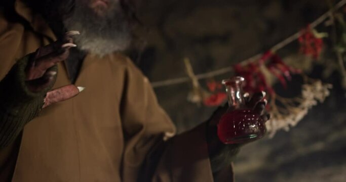 An old bearded sorcerer cooks in a cauldron near a medieval cave a magic potion of red color at night.