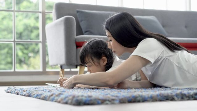 Cheerful Asian family spending time together in room.  Happy Asian mother and daughter lying on floor carpet drawing a picture or writing book with colour pencils at home. 