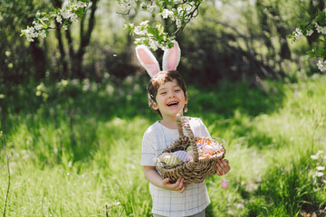 Easter egg hunt in spring garden. Funny boy with eggs basket and bunny ears on Easter egg hunt in...