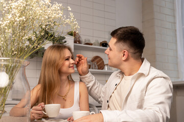 Obraz na płótnie Canvas Young couple sitting at kitchen table, holding cups with tea or coffee, looking at each other and talking. Happy wife and husband. High quality photo