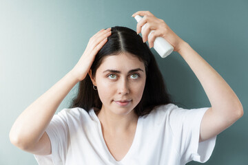 Portrait of a caucasian woman with dark hair applying spraying cosmetic product to the hair parting. Green background. The concept of hair care and dandruff problems