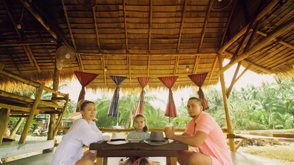 Fototapeta na wymiar The happy family eat and drink tasty beverages spending time in local floating cafe on water. Father, mother and daughter having breakfast outdoors. Man, woman and a child in a cafe. Tropical view