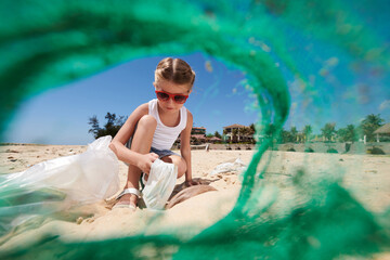 Little girl in sunglasses collecting garbage on sandy beach, view through plastic net