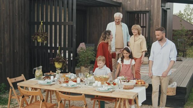 Wide shot of big Caucasian family of three generations walking to table with food on it in front yard of summer house on warm day, talking and smiling