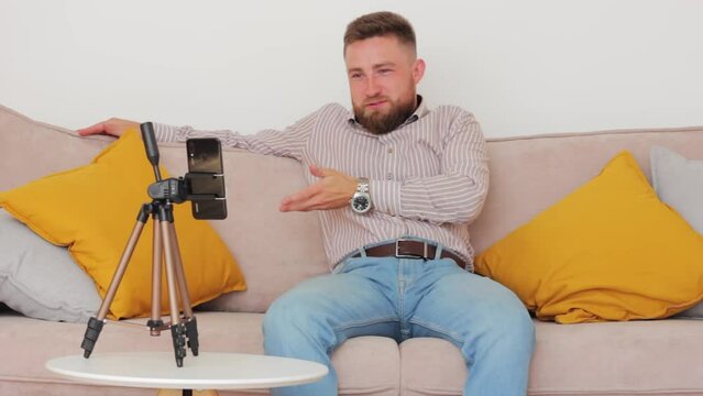 Caucasian unshaven blogger in shirt and jeans tells news about new high tech innovations holds mobile phone on tripod, looks at camera and gestures with his hands while sitting on dewan in room
