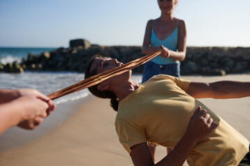 Young man bending backwards and passing under ribbon when ing limbo game with friends on beach - obrazy, fototapety, plakaty