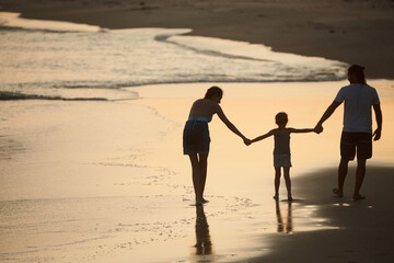 Happy family of three holding hands when walking on sandy beach at sunset