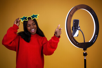 laughing African-American blogger girl in a red hoodie and big star glasses with a lighted ring lamp and phone pointing her fingers at it on a yellow background with copy space