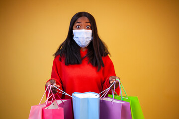 A surprised young African-American girl in a red sweatshirt and virus protection mask looking into open colorful packages on a yellow background with copy space