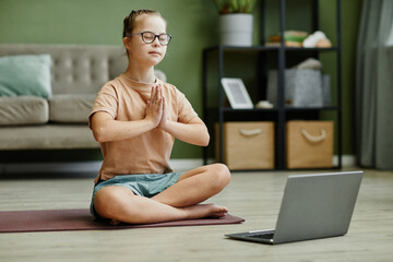Full length portrait of cute girl with down syndrome doing yoga at home, sitting on mat in lotus...