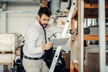 A focused graphic engineer on ladders in stockroom at printing shop holding sheet.