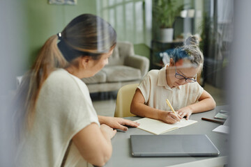 Portrait of young girl with down syndrome studying at home with female tutor helping, behind glass...