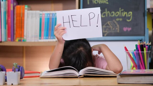 Asian little girl feeling sad and tired sitting at the table with many books and holding a paper with word Help. Learning difficulties. Home schooling.
