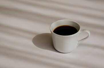 A cup of black coffee puts on white bed sheet with shadow of curtain in the morning. Relaxing at home concept.