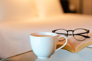 Selective focus of a white cup coffee puts on glass table with book and spectacles in bedroom. Relaxing at home concept.